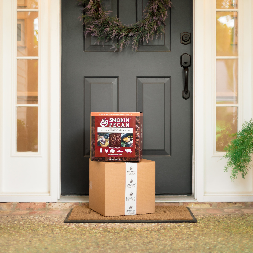 A 20 lb box of Smokin' Pecan Pecan Shell Pellets sits on a cardboard package on a doormat in front of a black door with a wreath.