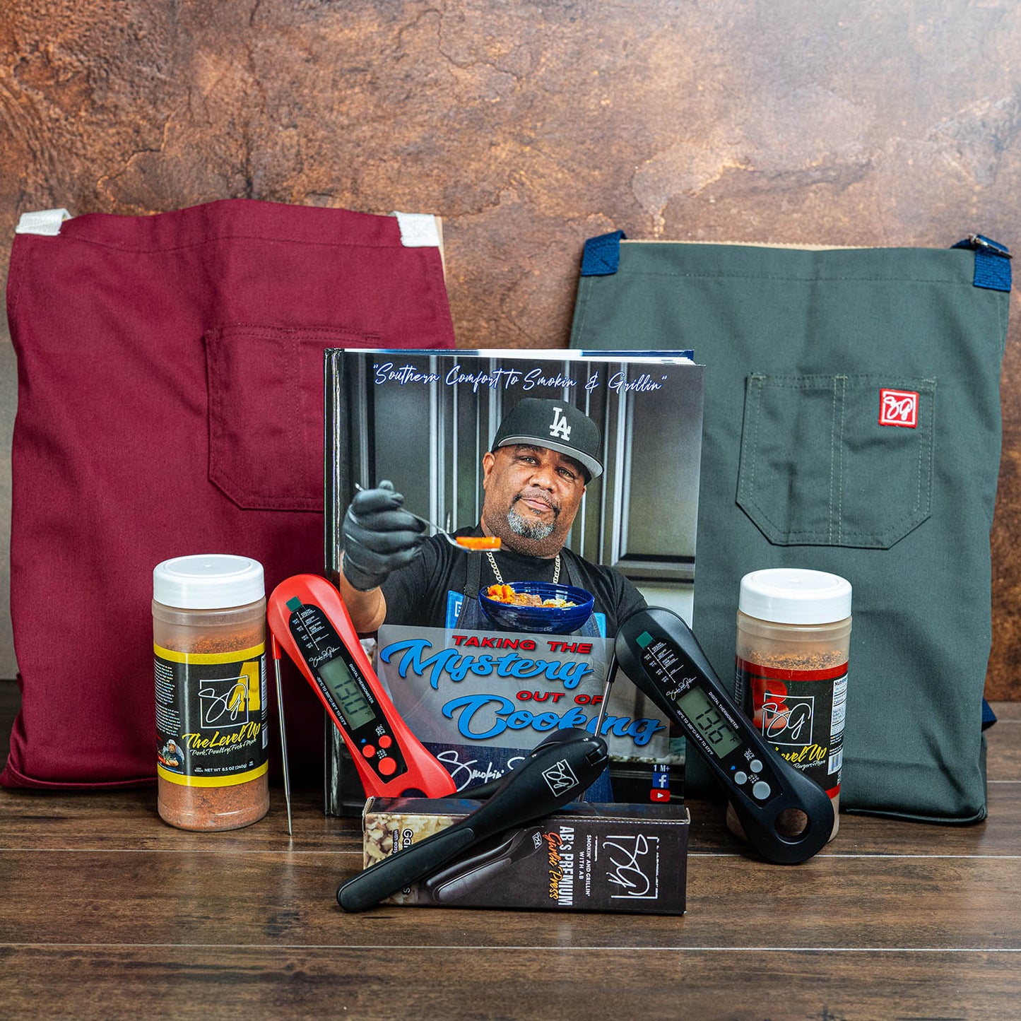 The Complete Kitchen Starter Bundle by Smokin and Grillin wit AB features a cookbook, maroon and dark green aprons, two digital meat thermometers, a seasoning duo, and a pen—all showcased on a wooden surface.