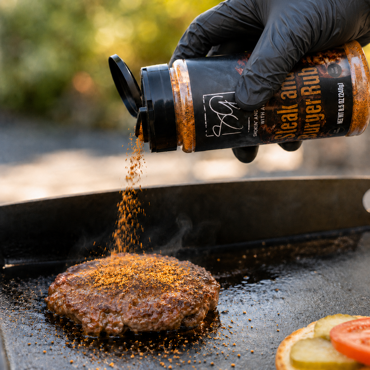 A gloved hand sprinkles Smokin and Grillin with AB’s steak and burger seasoning onto a burger on the grill—sliced tomato, onion, and pickle nearby. Perfect for flat-top cooking with AB’s Griddle Kit from Smokin and Grillin with AB.