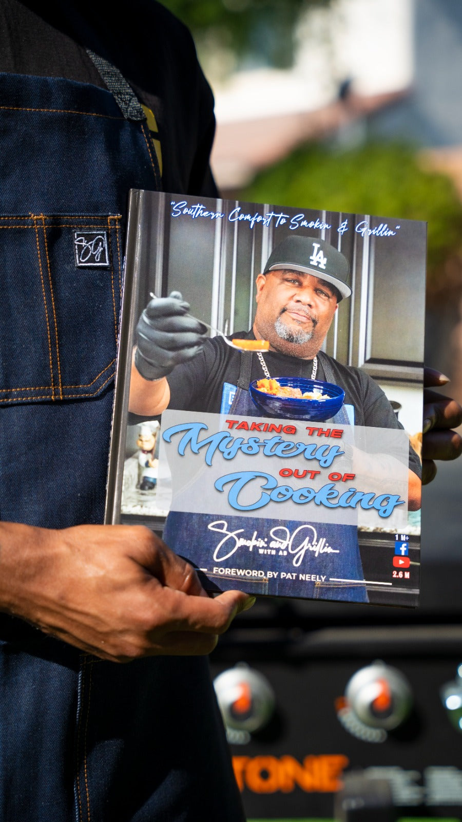 Someone in a dark apron holds up the Taking the Mystery out of Cooking Cookbook by Smokin and Grillin wit AB, featuring an approachable chef on the cover. Blurry grill knobs in the background hint at effortless, tasty meals ahead.