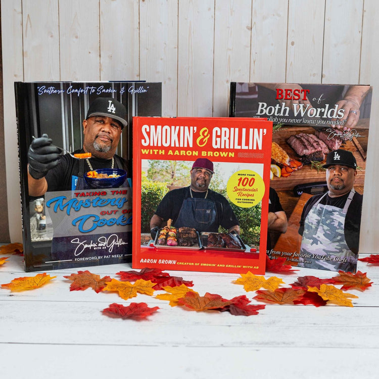 Three BBQ cookbooks by Aaron Brown are displayed upright on a wooden surface with autumn leaves scattered around. The center book is titled Smokin & Grillin with Aaron Brown.