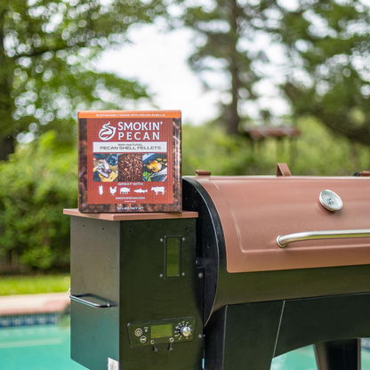 A 20 lb box of Smokin' Pecan Pecan Shell Pellets sits on a black and brown grill by a backyard pool, showcasing this eco-friendly barbecue fuel.
