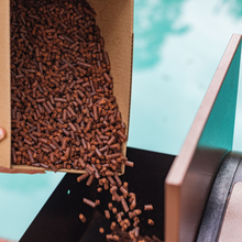 A hand pours Smokin' Pecan Pecan Shell Pellets from the 20 lb box into a pellet grill hopper, with a blurred blue background.