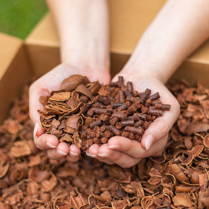 A pair of hands holds coarse pecan shells and Smokin’ Pecan Shell Pellets—2 lb Bag—in dark brown, above a box filled with similar shells.