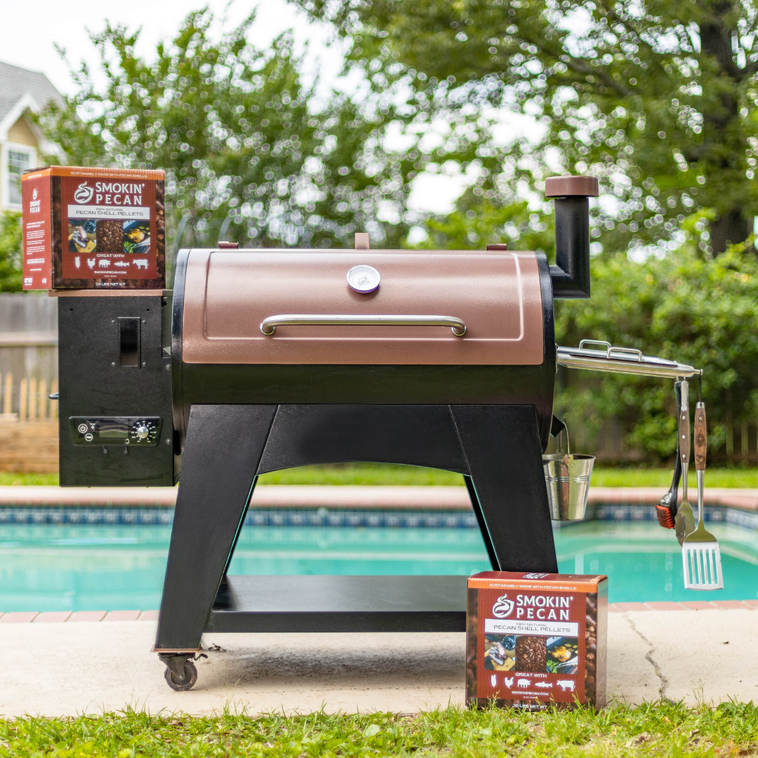 A pellet grill stands poolside with grilling utensils on the right and a 20 lb box of Smokin' Pecan Pecan Shell Pellets on each side. Trees and a fenced yard can be seen in the background.