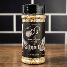 A plastic shaker bottle of AB's SPG (Salt, Pepper, and Garlic) by Smokin and Grillin wit AB sits on a wooden surface, featuring a black label against a backdrop of black and white tiles.
