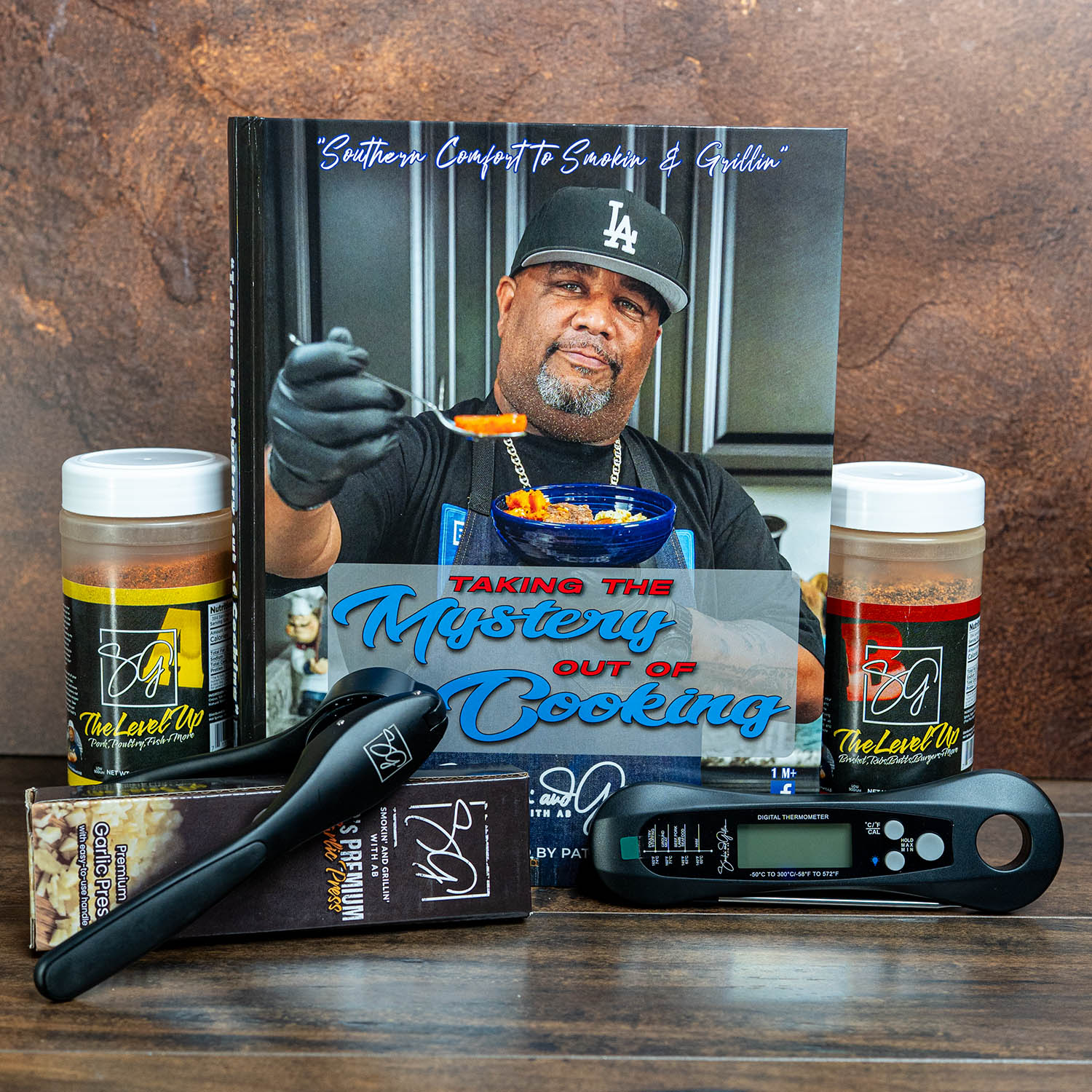 The Smokin and Grillin wit AB Kitchen Essentials Basics Bundle features the cookbook Taking the Mystery Out of Cooking, two seasoning jars, a meat thermometer, and a flashlight. The cover shows a man in a black LA cap holding a spoonful of food.