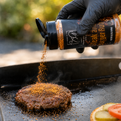 A gloved hand sprinkles Smokin and Grillin with AB’s steak and burger seasoning onto a burger on the grill—sliced tomato, onion, and pickle nearby. Perfect for flat-top cooking with AB’s Griddle Kit from Smokin and Grillin with AB.