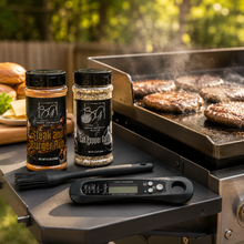 Smokin and Grillin with AB’s Griddle Kit—featuring seasoning bottles, a basting brush, and a digital meat thermometer—sits on a table by the grill with burger patties cooking, plus buns, cheese, and steak & burger seasoning in view.