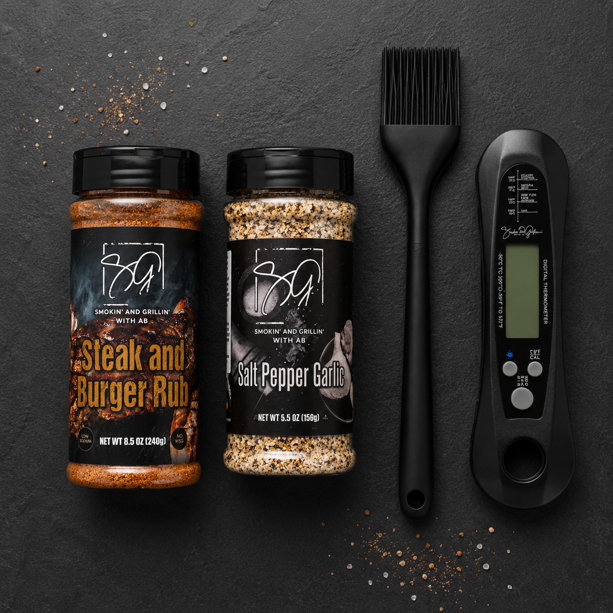Smokin and Grillin with AB's Griddle Kit features Steak and Burger Rub, Salt Pepper Garlic seasoning, a black basting brush, and a digital meat thermometer—perfect for flat-top cooking—shown on a dark surface sprinkled with seasonings.