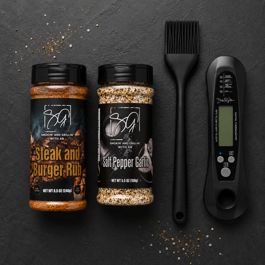Smokin and Grillin with AB's Griddle Kit features Steak and Burger Rub, Salt Pepper Garlic seasoning, a black basting brush, and a digital meat thermometer—perfect for flat-top cooking—shown on a dark surface sprinkled with seasonings.