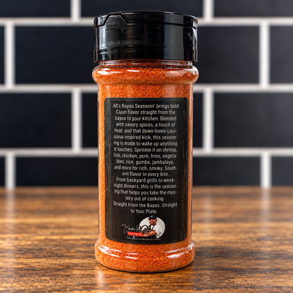 A plastic spice jar of AB’s Bayou Seasonin’ filled with reddish-orange seasoning, featuring a black label with flavor profile text and a small circular photo of a man in a chef’s coat.