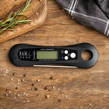 The SnG Food Thermometer by Smokin and Grillin wit AB, featuring a waterproof black design and grilling temperature guidelines, sits on a wooden surface next to rosemary, a wooden board, gray cloth, and peppercorns.