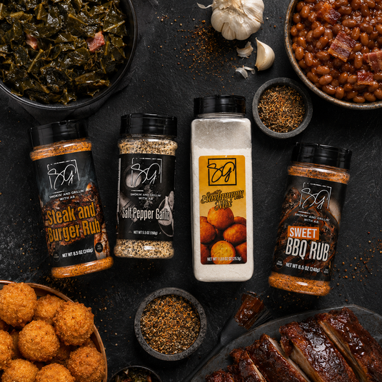 Four bottles from the Smokin and Grillin with AB Sunday Dinner Starter Pack sit on a dark surface, surrounded by collard greens, baked beans, ribs, hush puppies, and spices—the ultimate setup for unforgettable family meals.