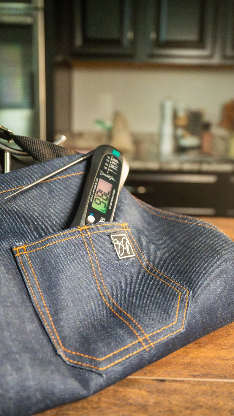 A Food Thermometer showing 92.2°F sits in a Denim Apron pocket on a wooden counter, with dark kitchen cabinets behind—showcasing the Smokin and Grillin wit AB Kitchen Bundle.
