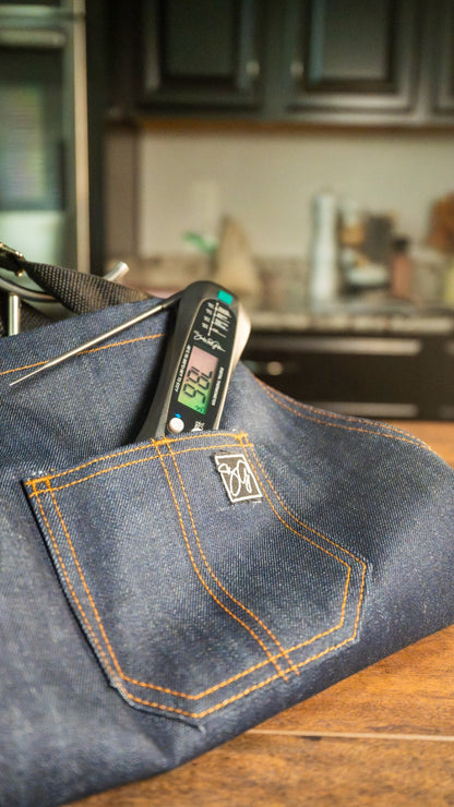 A Food Thermometer showing 92.2°F sits in a Denim Apron pocket on a wooden counter, with dark kitchen cabinets behind—showcasing the Smokin and Grillin wit AB Kitchen Bundle.