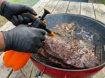 Wearing black gloves, a person sprays liquid from the Smokin and Grillin wit AB BBQ Spritz Bottle onto meat cooking on a grill with wood chips, using this essential tool as smoke rises from the grill.